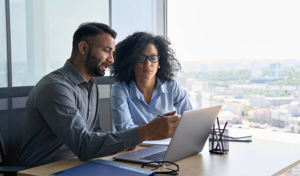 Multiethnic colleagues sitting at desk looking at laptop computer in office. Indian male ceo executive manager mentor giving consultation on financial operations to female African American colleague intern using laptop sitting in modern office near panoramic window. pointer stock pictures, royalty-free photos & images