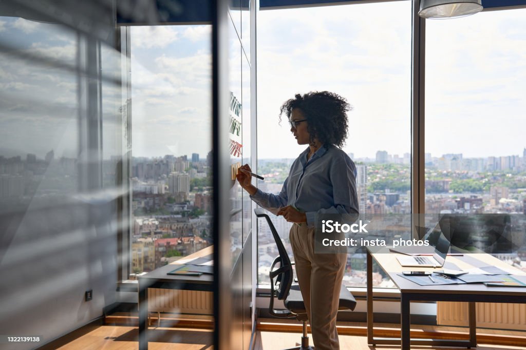 Afro American ceo writing on sticky notes in office with big city urban view. Young African american executive manager businesswoman writing strategy ideas on sticky notes on whiteboard doing business project presentation in contemporary corporate office with panoramic view. Whiteboard - Visual Aid Stock Photo