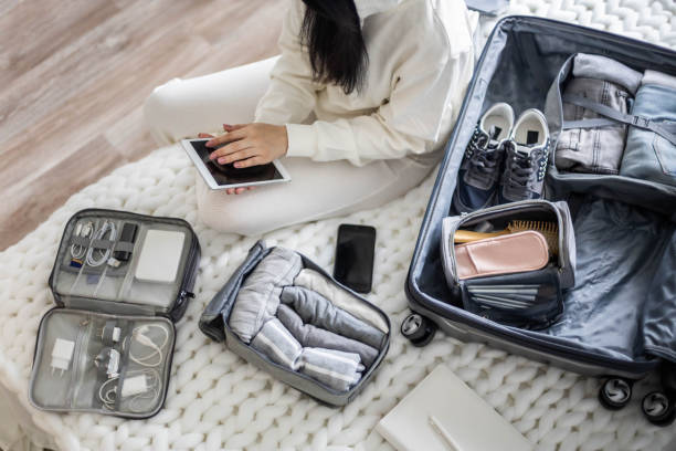 a beautiful young girl in a white casual suit sits on the bed and uses a tablet - suitcase imagens e fotografias de stock