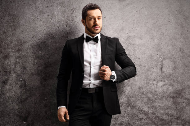 istock Young man in a black suit and bow tie leaning on a rusty gray wall 1322149115