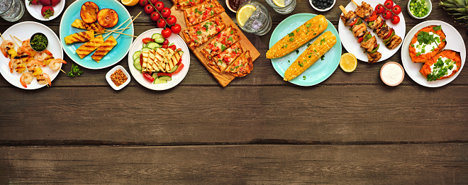 Summer BBQ grill top border over a dark wood banner background. Grilled flatbread, chicken and shrimp skewers, stuffed sweet potato, corn, fruit and salad. Above view with copy space.