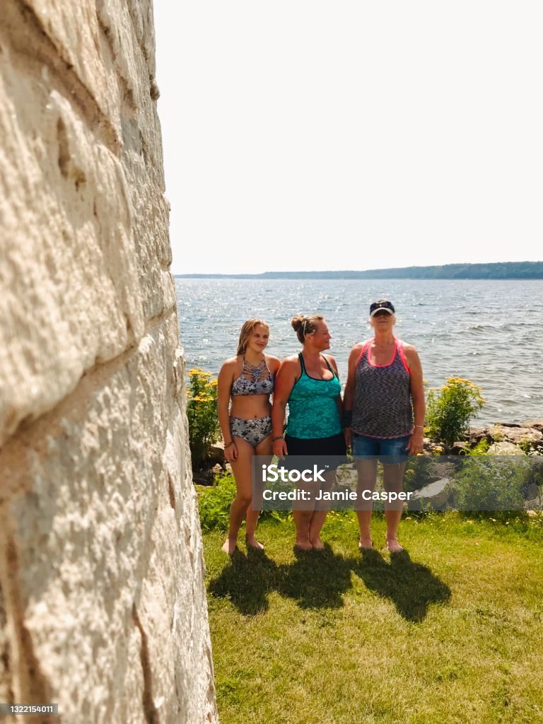 Three generations of female family members together on Lake Michigan. Three female family members, daughter, mother, and grandmother, standing outdoors near the lake shore and standing together showing three generations enjoying a day at the Lake. Partial view of lighthouse can be seen. Taken in Gladstone, Michigan in The Upper Peninsula. Adult Stock Photo