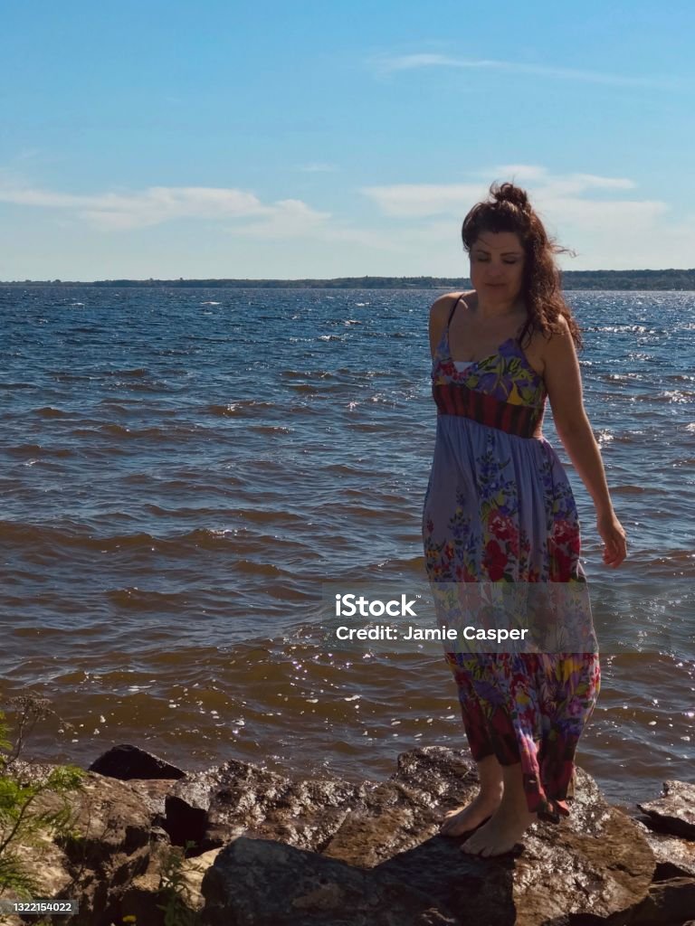 Summer dress on Lake Michigan. Photograph of beautiful mid aged woman, enjoying a lake breeze on the shores of Lake Michigan on a beautiful sun sparkling day while wearing a purple flowered sundress which is blowing around gently from the lake breeze. Fashion Stock Photo