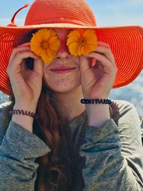 This is my summer face. Cute red head freckle faced teen girl at the beach, a head shot of her wearing a large orange straw beach hat and holding up matching orange flowers over her eyes and a wide cute smile on her face. gladstone michigan stock pictures, royalty-free photos & images