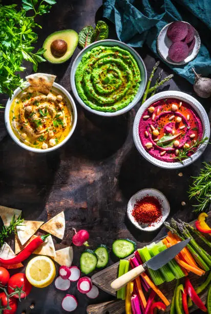 Photo of Hummus Three bowl of chickpeas, avocado and beetroot with cut vegetables sticks on dark wood table