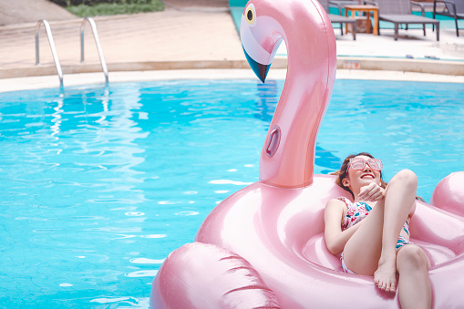 Happy traveler asian woman with bikini relax on big pink flamingo pool float in swimming pool at Thailand, summer travel vacation concept