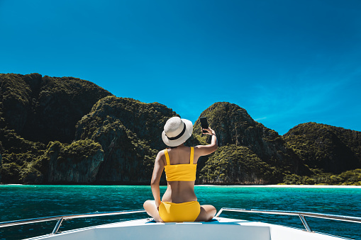 Travel summer vacation concept, Happy solo traveler asian woman with bikini and mobile phone relax and selfie on boat in Maya Bay Phuket Thailand