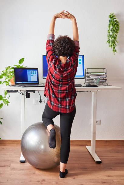 Woman stretches while working at an adjustable desk standing Woman stretches while working at an adjustable desk standing with one knee resting on a fitball standing desk photos stock pictures, royalty-free photos & images