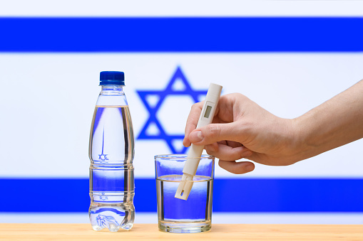 A hand with a water tester makes a measurement in a glass of clear water against the background of the flag of Israel. Test and assessment of drinking water supplies in Israel.
