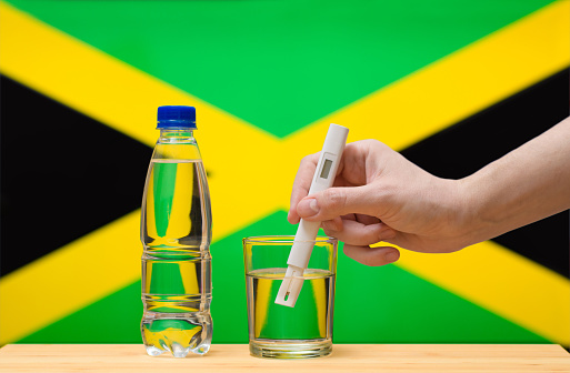 A hand with a water tester makes a measurement in a glass of clear water against the background of the flag of Jamaica. Test and assessment of drinking water supplies in Jamaica.