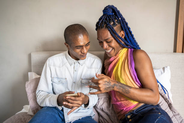 아프리카 계 미국인 비 바이너리 사람과 트랜스 젠더 여성은 스마트 폰과 대화를 사용하여 침대에서. 성소수자 부부. - non cisgender 뉴스 사진 이미지