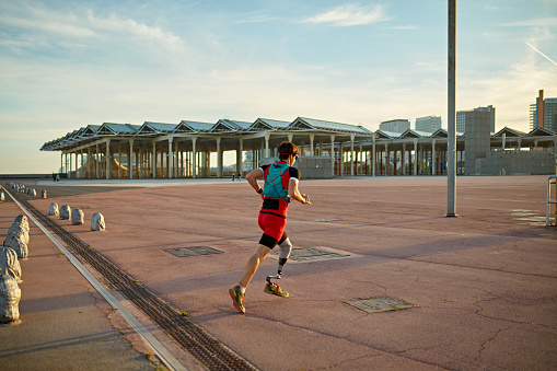 Full length view of early 30s Caucasian male athlete in hydration vest and sports attire moving past camera while exercising in Barcelona public park.