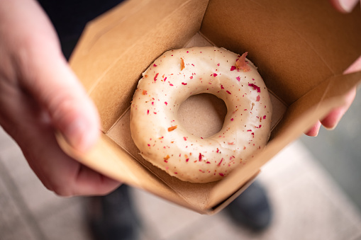 Naturally colored and flavored with Earl Grey tea..  Anonymous Asian man holding compostable takeout cardboard box with one vegan, organic, gluten-free cake donut inside.