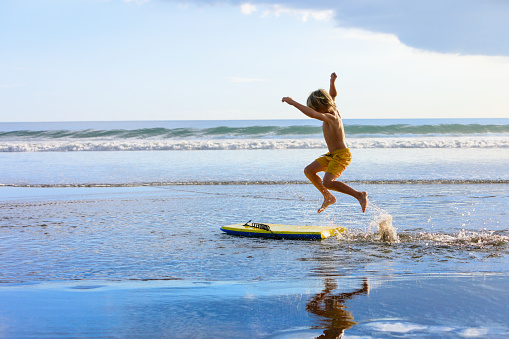 Happy boy - young surfer with bodyboard have fun on beach, run by sea water pool. Active family lifestyle, kids outdoor water sports, swimming activity in surf camp. Summer vacation with child.
