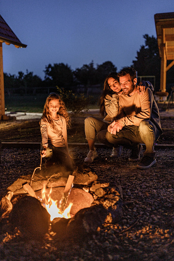 Happy parents and their small girl enjoying while spending a night by the campfire in the backyard.