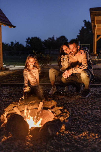 Happy family enjoying by the bonfire in the backyard at night. Happy parents and their small girl enjoying while spending a night by the campfire in the backyard. Bonfire stock pictures, royalty-free photos & images