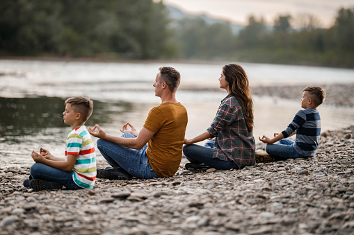 Parents and their kids exercising Yoga in Lotus position on a stone beach by the river. Copy space.