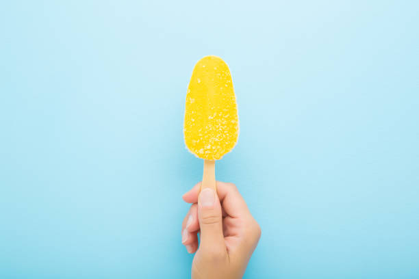 Young adult woman hand holding bright yellow mango ice cream with stick on light blue table background. Pastel color. Closeup. Top down view.
