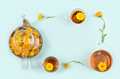 Frame made from cups of herbal tea, transparent teapot and calendula flowers on blue background. Calming drink concept. Copy space Top view Flat lay