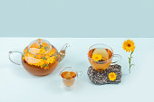 Cups of herbal tea, transparent teapot with calendula flowers and stones on blue background. Calming drink concept