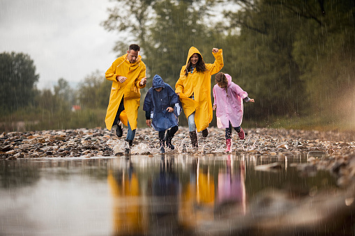 Happy family in raincoats having fun while stepping into big puddle during rainy day in nature. Copy space.