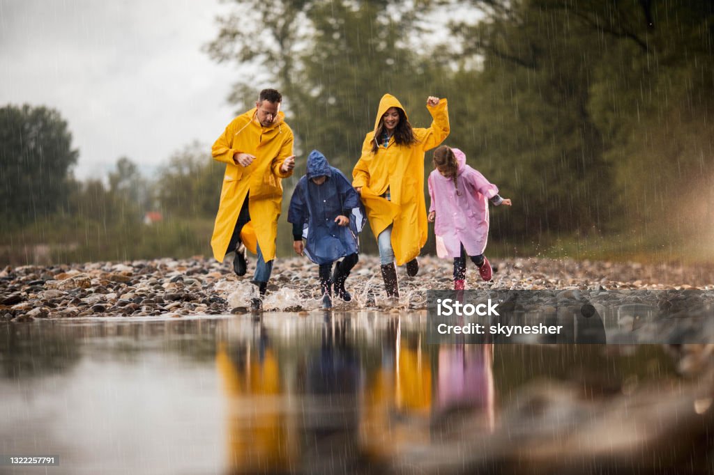 Carefree family having fun while running into water during rainy day. Happy family in raincoats having fun while stepping into big puddle during rainy day in nature. Copy space. Rain Stock Photo