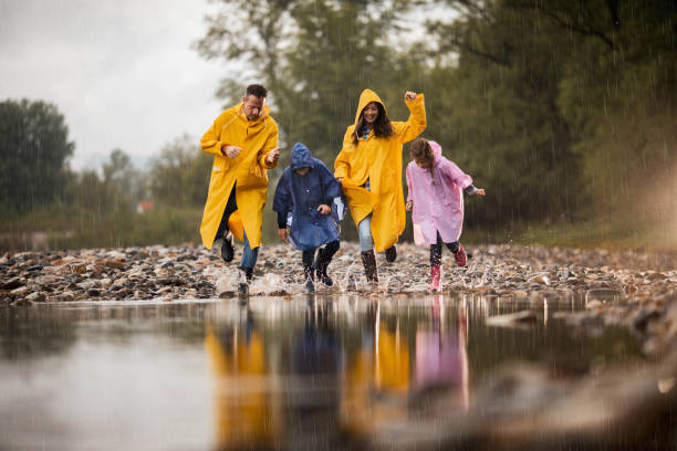 Carefree family having fun while running into water during rainy day. Happy family in raincoats having fun while stepping into big puddle during rainy day in nature. Copy space. spring flowing water stock pictures, royalty-free photos & images