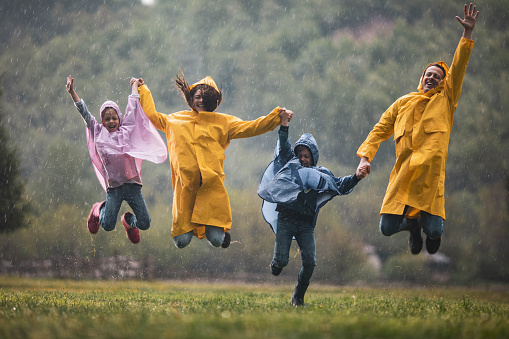 Happy family in raincoats having fun while holding hands and jumping together during rainy day in nature. Copy space.