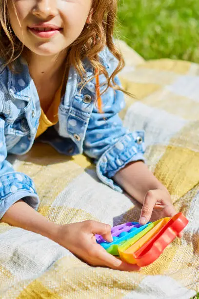 Photo of Girl lies on a blanket outdoors and play pop it, kid hands playing with colorful pop It