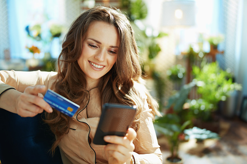 Green Home. happy trendy middle aged woman with long wavy hair with credit card making online shopping on e-commerce website in the modern house in sunny day.