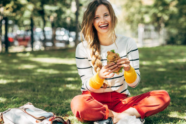 Happy student female sitting on the green grass at the college campus on a sunny day, have lunch and studying outdoors. A smiling young woman takes a rest eating fast food and learning in the park. stock photo