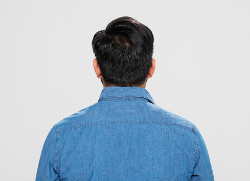 Rear view of young man in denim shirt standing against grey background.