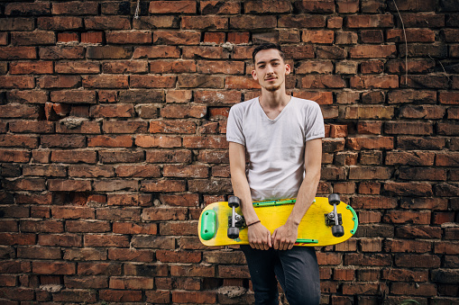 One man, portrait of a young man standing by a brick wall. He is holding skateboard.