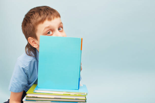 istock Schoolboy is sitting with a stack of books and reading and covers his face with a book isolated on blue background 1322278858