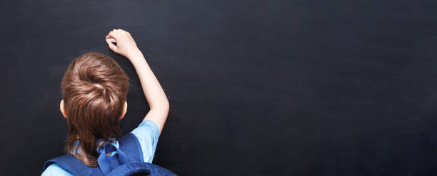 istock Boy standing back in front of school blackboard and writing. banner 1322279136