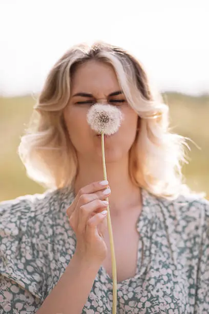 Photo of young woman hid behind a dandelion and grimaced funny. Funny photo. Pollen allergy concept