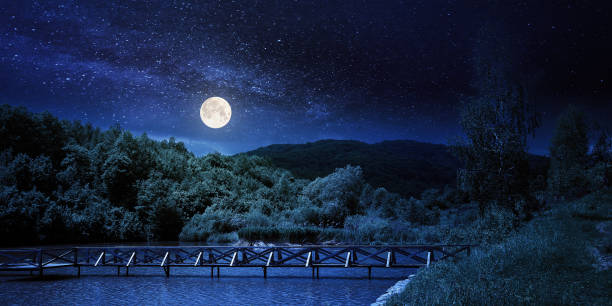 lake among mountain landscape in spring at night. beautiful countryside scenery with forest on the shore in full moon light. clouds on the sky