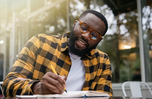 Portrait of happy African American business man writing on notebook, working project. Smiling university student studying, exam preparation, taking notes. Education concept