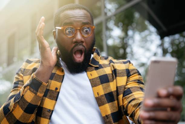 istock Excited young man with open mouth using mobile phone, looking at digital screen, sitting at workplace. Portrait of emotional African American freelancer missed deadline 1322285883
