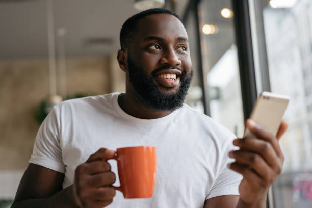 Handsome African American man using mobile phone, drinking coffee in cafe. Freelancer holding smartphone, working from home