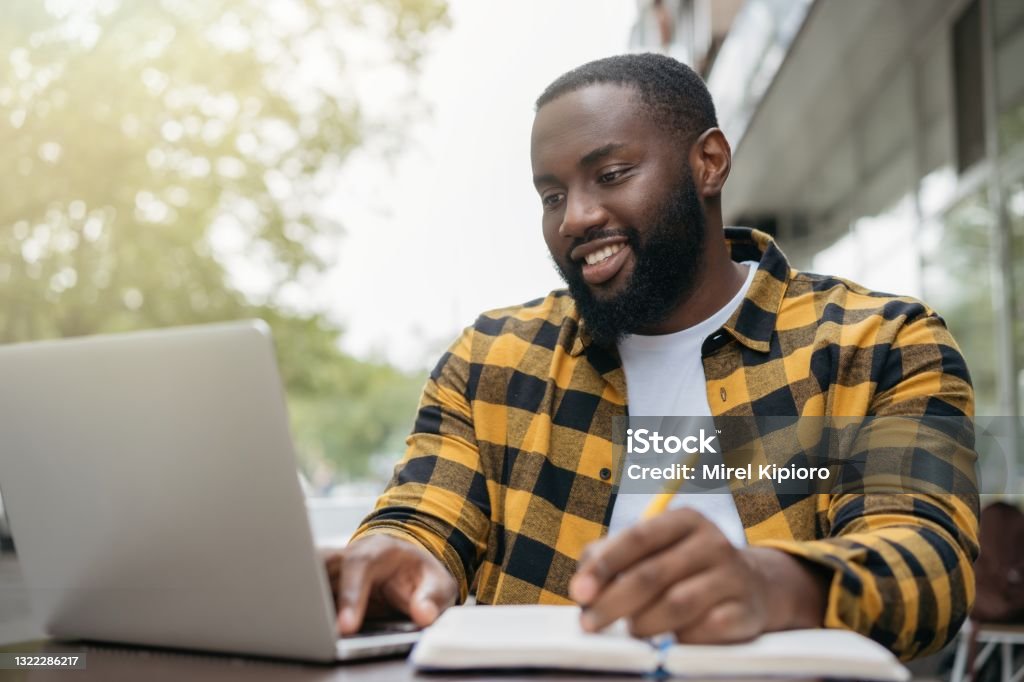 African American man using laptop computer, taking notes, planning start up, working online. Portrait of happy student studying, learning languages, online education concept Young handsome African American man using laptop computer, taking notes, planning start up, working online. Portrait of happy student studying, learning languages, online education concept E-Learning Stock Photo