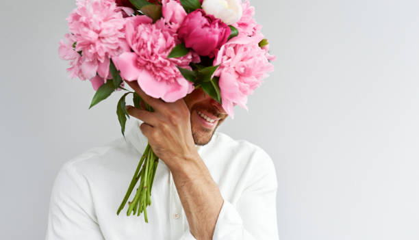 Closeup of handsome man covers his eyes with the bouquet of pink peonies as a gift for Valentine's day or wedding day. Smiling male carrying flowers in the hands, isolated on the grey background. stock photo