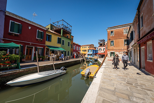 Burano, Italy - June 2th, 2021: Burano island with multi colored houses in front of a small canal with moored boats, restaurants and shops. Venice lagoon, UNESCO world heritage site, Veneto, Italy, Europe. A group of tourists visit the famous island on a sunny spring day with clear sky.
