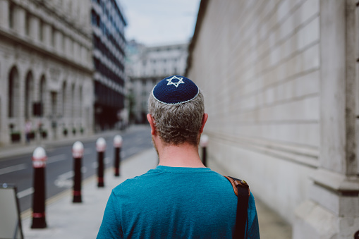 Rear view image depicting a mid adult Jewish man in his 30s wearing a traditional Jewish skull cap (with star of David design). He is walking on a street in central London, surrounded by city architecture.