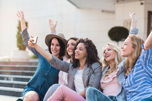 Fashionable attractive young women sitting on the stairs in the street, taking a selfie