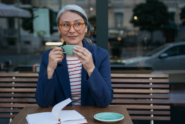 카페에 앉아 커피를 마시는 성숙한 사업가. 세련된 안경을 쓰고 차 한 잔을 들고 있는 펜서티 아시안 여성의 초상화 - pensive women business mature adult 뉴스 사진 이미지