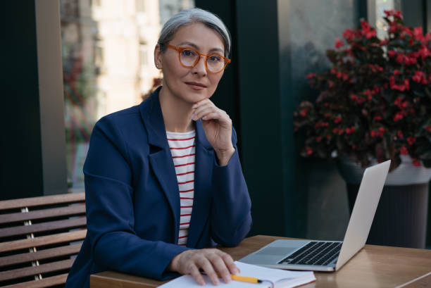 nachdenkliche reife geschäftsfrau, die in die kamera schaut und am arbeitsplatz sitzt. zuverlässiger freelancer arbeitet, mit laptop, planungsprojekt - women serious mature adult business stock-fotos und bilder