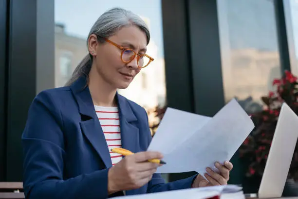 Photo of Mature businesswoman reading contract, planning project, brainstorming. Portrait of attractive asian secretary reading, working with documents, sitting at workplace