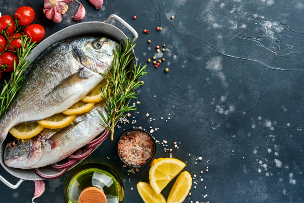 istock Raw dorado fresh fish or sea bream with ingredients for making lemon, thyme, garlic, cherry tomato and salt on a black slate, stone or concrete background. Top view with copy space. 1322309368