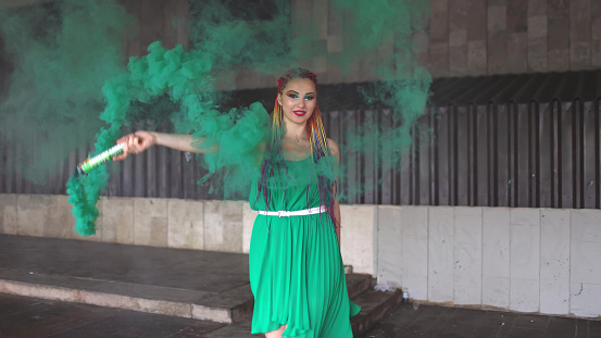 A young cheerful girl in a bright green spring dress with makeup with sequins and rainbow African braids. She stands near a building and blows green smoke in the street. UHD 4K 4x slow-motion video